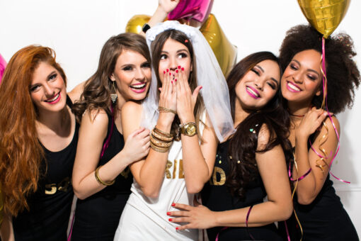 Young girlfriends celebrating bride night. Bride is wearing a veil and a T-shirt with the inscription ''BRIDE'', bridesmaids are wearing T-shirt with the inscription ''Squad''. They are laughing, dancing and singing. Room is decorated with star and heart shaped balloons.